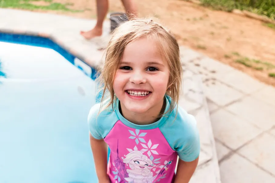 A smiling little girl in a swim shirt beside a sparkling pool